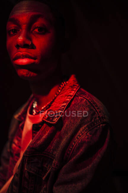 Crop calm stylish African American man in jeans jacket under neon red light in shade on black background looking at camera — Stock Photo