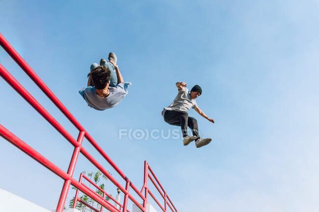 From below fearless male friends jumping above metal railing in city while performing parkour stunt on sunny day — Stock Photo