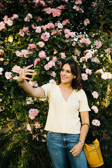 Fröhliche junge Frau meldet sich per Handy, während sie im Frühlingsgarten vor einem blühenden Strauch mit rosa Blüten steht — Stockfoto