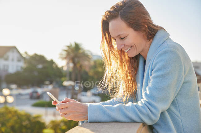 Улыбающаяся взрослая женщина в теплом пальто просматривает телефон, опираясь на забор возле городской улицы в солнечный день — стоковое фото