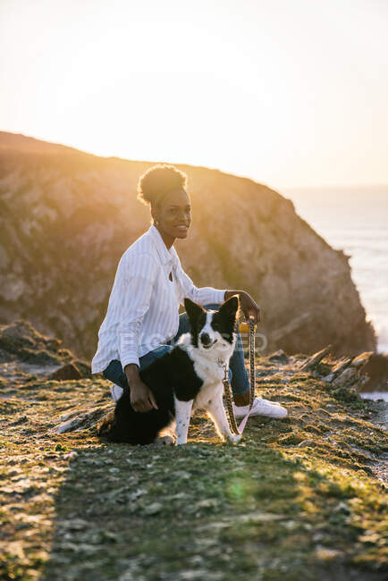 Vista lateral de la joven propietaria afroamericana con perro Border Collie pasando tiempo juntos en la playa cerca de ondear el mar al atardecer contemplando vistas - foto de stock