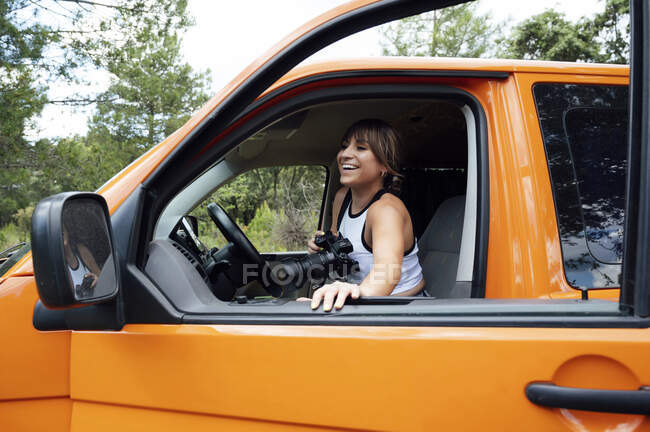 Vista lateral de la alegre viajera sentada en furgoneta con cámara fotográfica durante las vacaciones de verano en la naturaleza - foto de stock