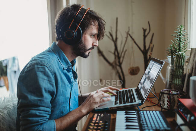 Vista lateral do jovem em fones de ouvido usando sintetizador e laptop na mesa em casa — Fotografia de Stock