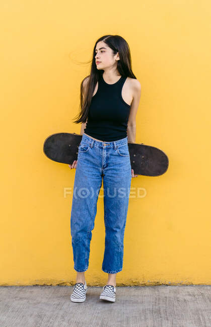 Young female skater with skateboard standing looking away on walkway with colorful yellow wall on the background in daytime — Stock Photo