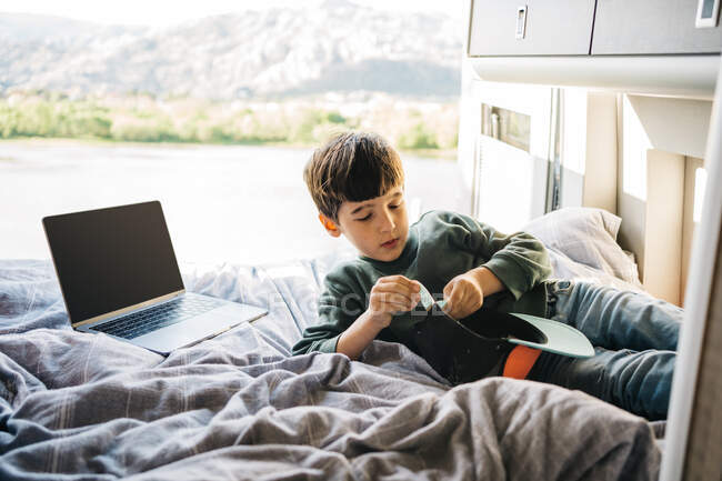 Маленький хлопчик розслабляється всередині автономного будинку, лежачи на ліжку поруч з ноутбуком і шапочкою — стокове фото