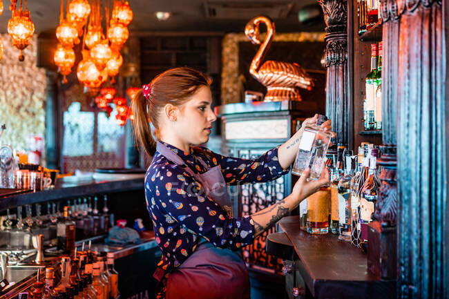 Side view of female bartender choosing alcohol drink in bottle for preparing cocktail in bar — Stock Photo