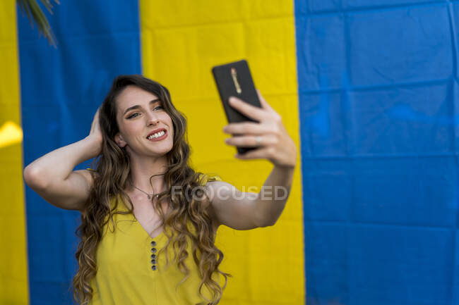Mujer con el pelo ondulado tomar selfie con el teléfono móvil mientras se ríe en dos fondo de color en la calle - foto de stock