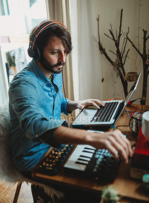 Vista laterale del giovane in cuffia con sintetizzatore e laptop a tavola a casa — Foto stock