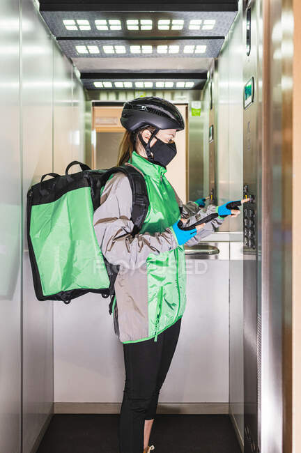 Female courier in uniform and mask using smartphone and pushing button in elevator while making delivery during pandemic — Stock Photo