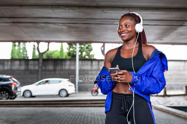 Sorridente giovane donna atletica afro-americana che ascolta musica nel parcheggio — Foto stock