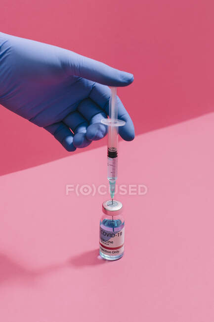 Обрезанная неузнаваемая рука врача, держащая шприц с иглой, готовящейся к коронавирусной вакцинации во фляжке на розовом фоне — стоковое фото
