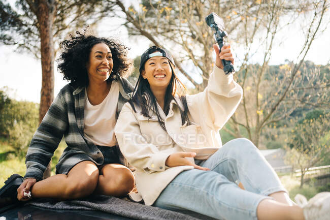 Low angle of cheerful young multiracial girlfriends sitting on roof of camper van and taking pictures while spending summer holidays together in countryside — Stock Photo