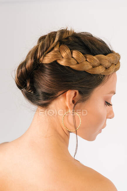 Side view of beautiful young female model with elegant braided hairstyle and makeup and with golden hoop earrings against white background — Stock Photo