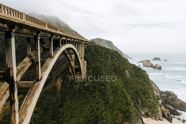 Breathtaking landscape of amazing white arched Bixby Bridge over river in deep valley against sloping mountains covered lush green grass and wildflowers in Big Sur in USA — Stock Photo