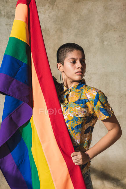 Serieux jeune femme bisexuelle ethnique avec drapeau multicolore représentant les symboles LGBTQ et détournant les yeux le jour ensoleillé — Photo de stock