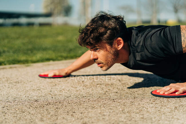 Ground level of attentive male athlete in plank pose with gliding discs training and looking forward on pavement in town — Stock Photo