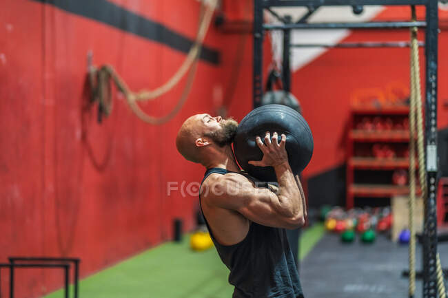 Вид сбоку сильный спортсмен поднимает тяжелый мяч во время тренировки в современном тренажерном зале — стоковое фото
