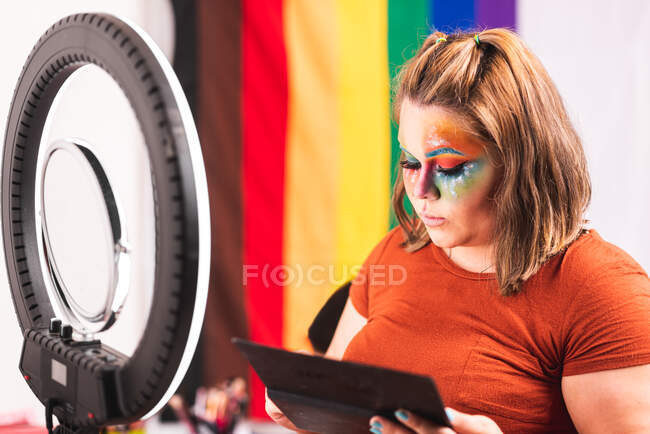 Плюс розмір жінки, яка дивиться на дзеркало з кільцевим світлом і застосування творчого макіяжу проти ЛГБТ прапора в студії — стокове фото