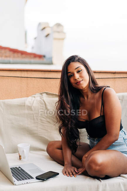 Young Hispanic female browsing on netbook while working remotely sitting cross legged on sofa with hot coffee mug in the morning on balcony — Stock Photo