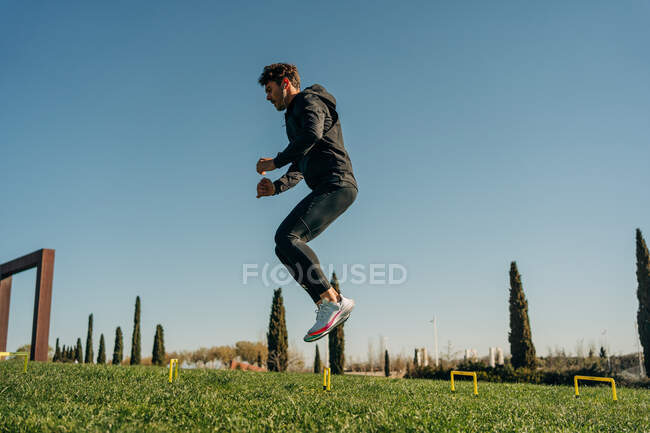 Vista laterale dell'atleta in forma maschile in abbigliamento sportivo che salta sul prato durante l'allenamento sotto il cielo blu — Foto stock