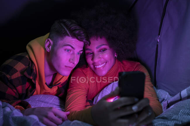 Happy multiracial man and woman smiling resting and browsing cellphone in tent at night — Stock Photo