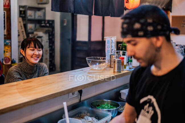 Азиатка в повседневной одежде сидит за стойкой и разговаривает с работником современного рамен-бара — стоковое фото