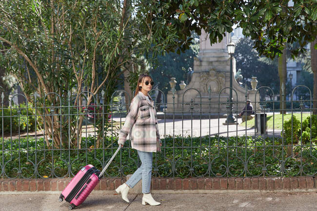 Вид всего тела сбоку на молодую женщину в стильном наряде и солнцезащитных очках, тянущую розовый чемодан во время прогулки по тротуарам города — стоковое фото