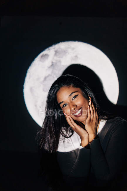 Joven mujer hispana alegre mirando a la cámara tocando mejillas mientras descansa bajo la proyección de la luna por la noche - foto de stock