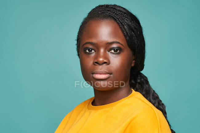 Unemotional regordeta afroamericana hembra en suéter amarillo de pie mirando a la cámara contra la pared azul - foto de stock