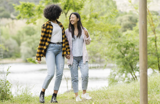 Content LBGT couple of multiracial females holding hands and walking along lake in woods — Stock Photo