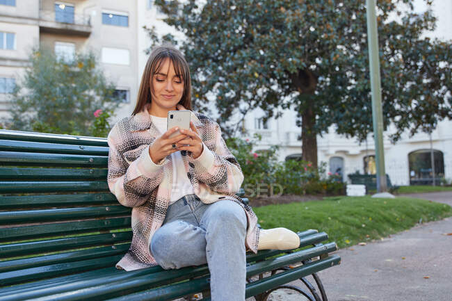 Сучасна тисячолітня жінка в стильному весняному вбранні сидить на лавці і переглядає мобільний телефон, відпочиваючи на міській вулиці, дивлячись далеко — стокове фото