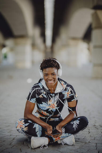 Young fitness African American woman resting and stretching legs while listening to music after running on city street — Stock Photo