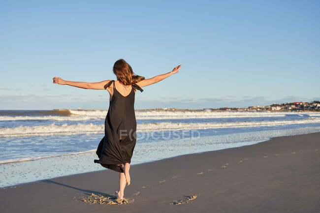 Полный вид сзади женщины в летнем платье, стоящей на песчаном берегу моря — стоковое фото