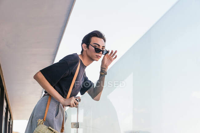 Baixo ângulo vista lateral do macho gay na moda óculos de sol apoiados em trilhos de vidro na varanda e olhando para longe — Fotografia de Stock