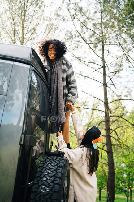 Side view of young African American woman helping friend climbing up camper van parked in green forest during summer adventure in countryside — Stock Photo