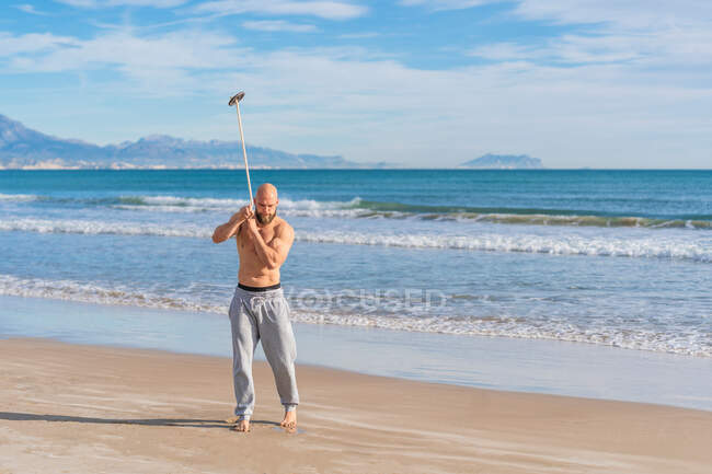 Full length of adult sportive man looking away doing around body pass with weight plate on band while warming up on sunny coast — Stock Photo