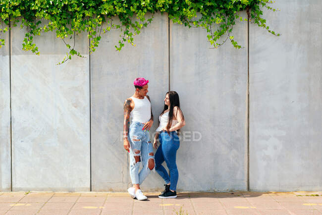 Contenidos homosexuales masculinos con el pelo rosa y la mujer amable de pie cerca de la pared en la ciudad y mirándose el uno al otro - foto de stock