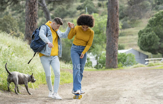 Full body young man holding hand and teaching cheerful black girlfriend to ride skateboard on countryside road near dog in nature — Stock Photo