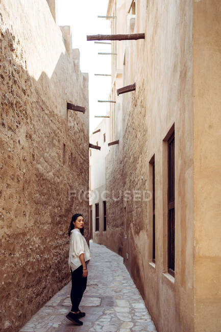 Vista trasera de una joven viajera vestida con ropa casual parada en un camino empedrado entre antiguos edificios de piedra mientras explora las calles del Barrio Histórico Al Fahidi en Dubái - foto de stock