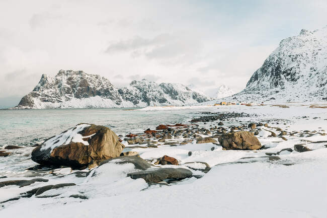 Boulders situado en la costa blanca nevada cerca del mar y las montañas en el día gris de invierno en las Islas Lofoten, Noruega - foto de stock