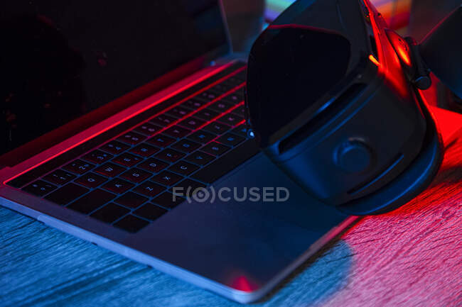 High angle of wooden desk with opened netbook near phone and VR goggles in dark room with neon lights — Stock Photo