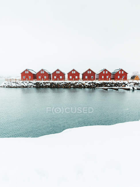 Cabañas rojas y muelle nevado situado cerca del mar ondulante en el frío día de invierno en la aldea costera en las Islas Lofoten, Noruega - foto de stock