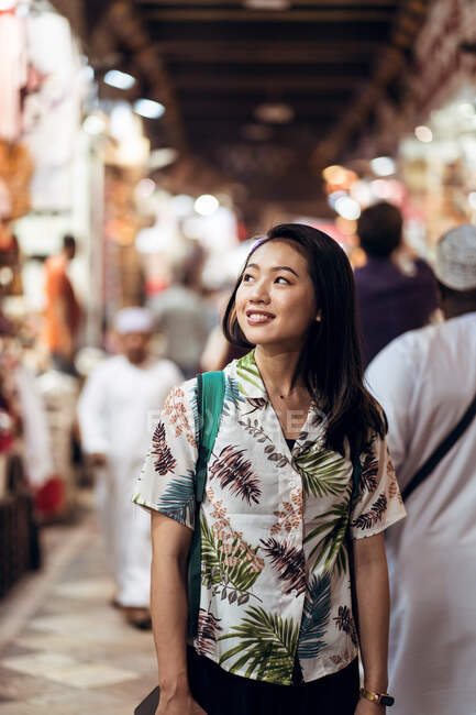 Happy Asian female traveler in tropical shirt with backpack looking away while standing on bazaar against blurred crowd in Doha — Stock Photo
