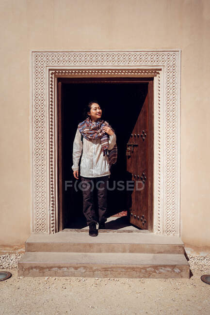 Encantada turista asiática en bufanda oriental tradicional sonriendo a la entrada de la vieja casa con adorno oriental y madera con puerta de decoración forjada en Doha - foto de stock