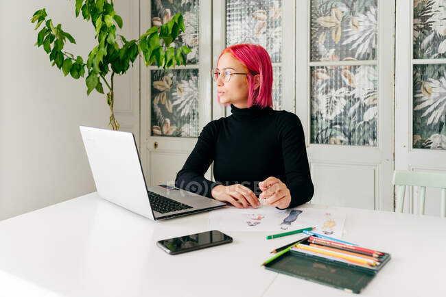 Молодая креативная женщина-дизайнер в повседневной одежде и очках задумчиво смотрит в сторону, сидя за столом с гаджетами и рисуя эскизы с красочными карандашами — стоковое фото