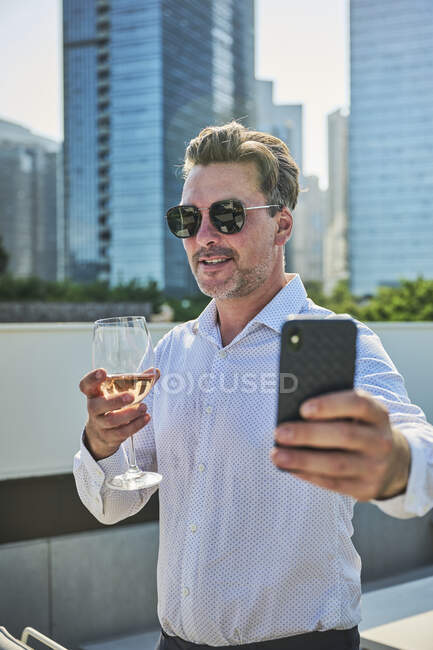 Designer après le travail ayant du temps de détente dans une terrasse à côté des immeubles de bureaux, il prend une tasse de vin blanc tout en prenant un selfie — Photo de stock