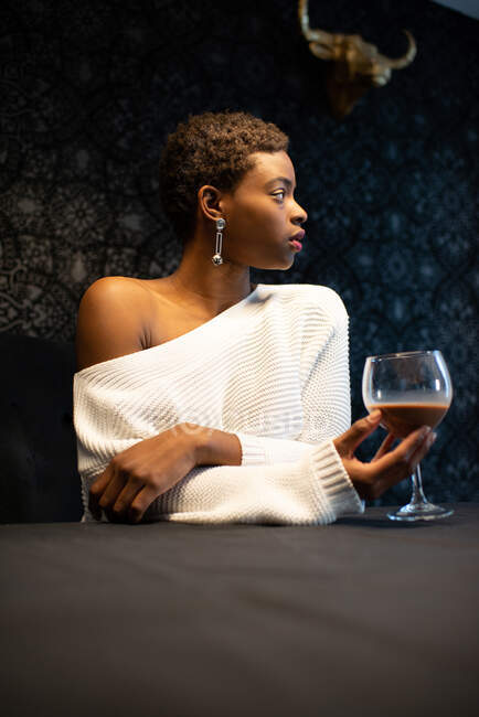 Mulher afro-americana com um copo de refrescante coquetel sentado à mesa e gelado no quarto escuro durante o fim de semana — Fotografia de Stock