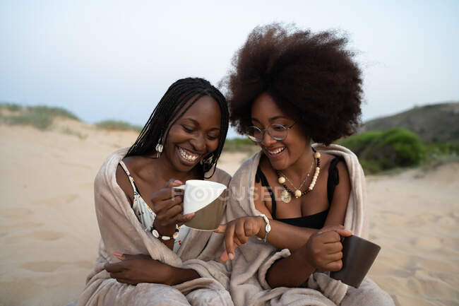 Feliz joven negro hembra mejores amigos con tazas de bebidas calientes sentado cerca envuelto en manta caliente y divertirse durante la noche de verano en la playa de arena - foto de stock