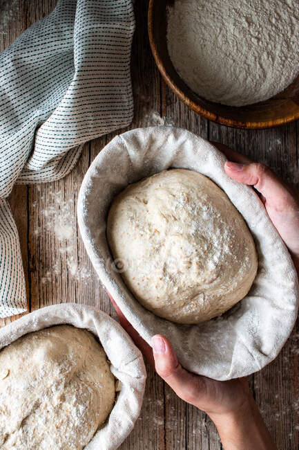 Mani di donna Dall'alto tenendo il pane di pasta madre su stand di vimini con stoffa su tavolo di legno con farina sparsa — Foto stock