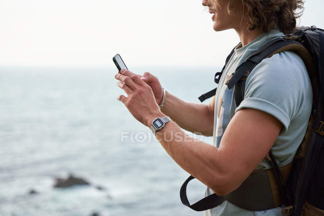 Вид сбоку мужчины-туриста, просматривающего смартфон, стоя на холме на фоне моря во время похода летом — стоковое фото